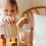 Cute little girl in white casual clothes standing near table and playing with wooden blocks while spending time at home