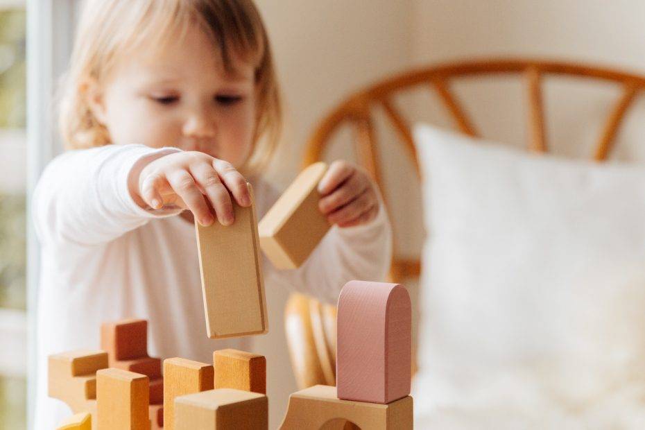 Cute little girl in white casual clothes standing near table and playing with wooden blocks while spending time at home